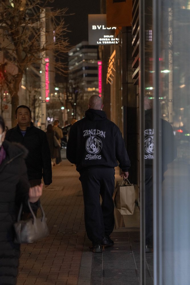 Hugh from behind in a Zen Den Studios hoodie walking along a Ginza street at night with the Bvlgari building visible — Zen Den Studios